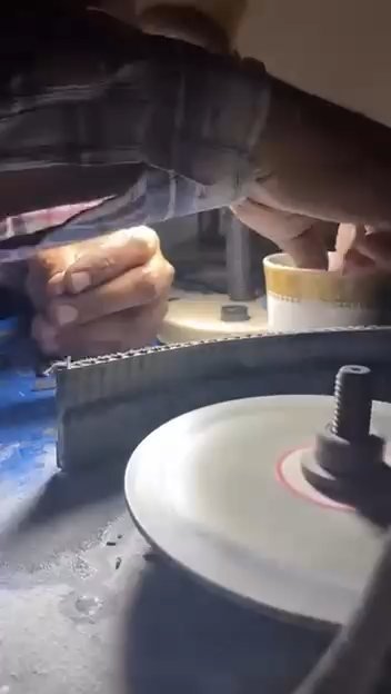 Hands holding a gemstone against a grinding wheel under a work lamp